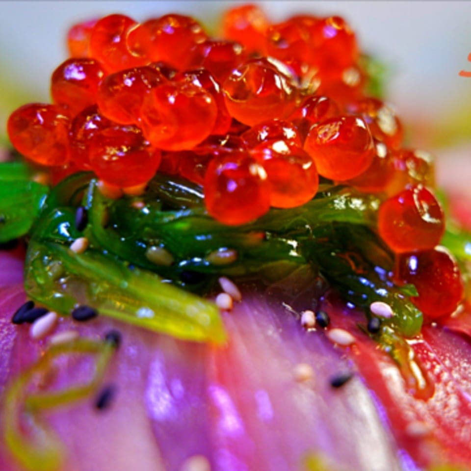 Close-up of vibrant red salmon roe and green seaweed salad on a bed of tuna sashimi.