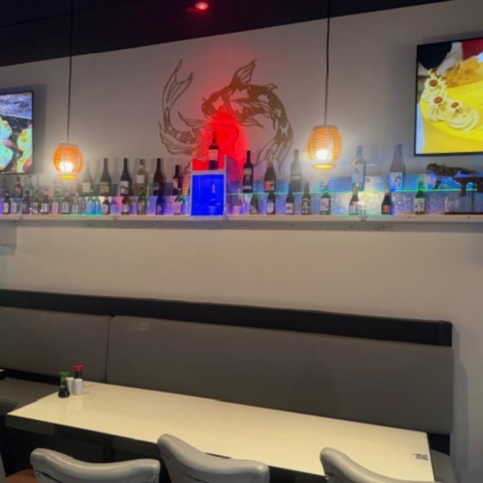 Interior of Sushi Yama restaurant with a bar shelf displaying bottles, illuminated by colorful lights and hanging lanterns.