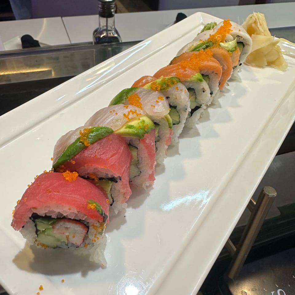 A long white platter holds a colorful sushi roll with tuna, salmon, avocado, and roe.