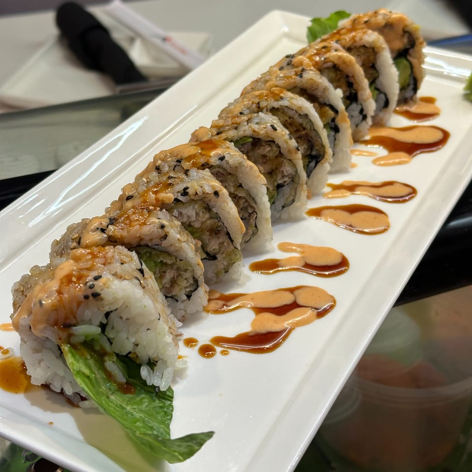 A row of Japanese sushi rolls with creamy sauce and sesame seeds on a white rectangular plate.