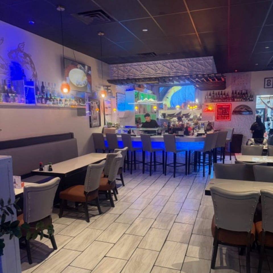 Interior of Sushi Yama restaurant with a blue-lit bar, dining tables, and a sushi chef.