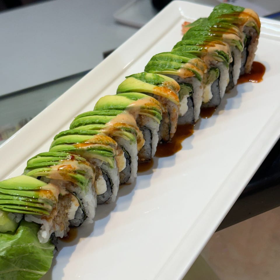 A long sushi roll with avocado slices and sauce on a white rectangular plate at Sushi Yama.