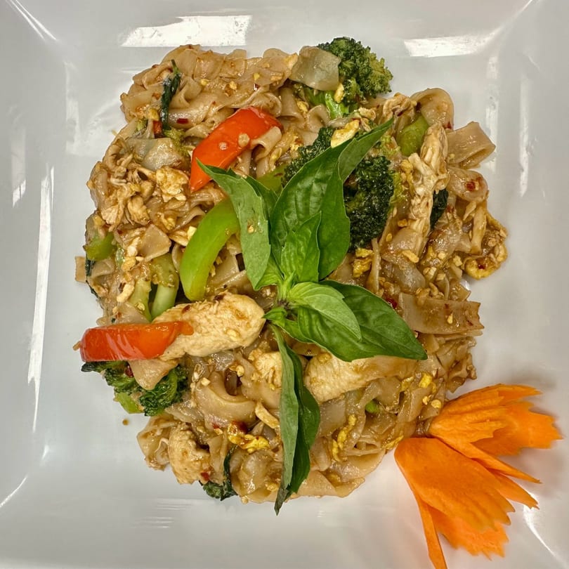 Pad See Ew with chicken, broccoli, red peppers, and basil, garnished with a carrot flower.